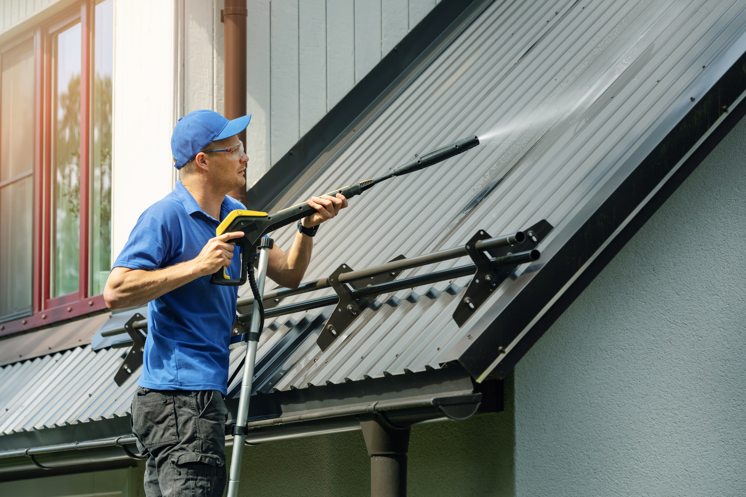 Man standing on ladder and cleaning house metal roof with high pressure washer.